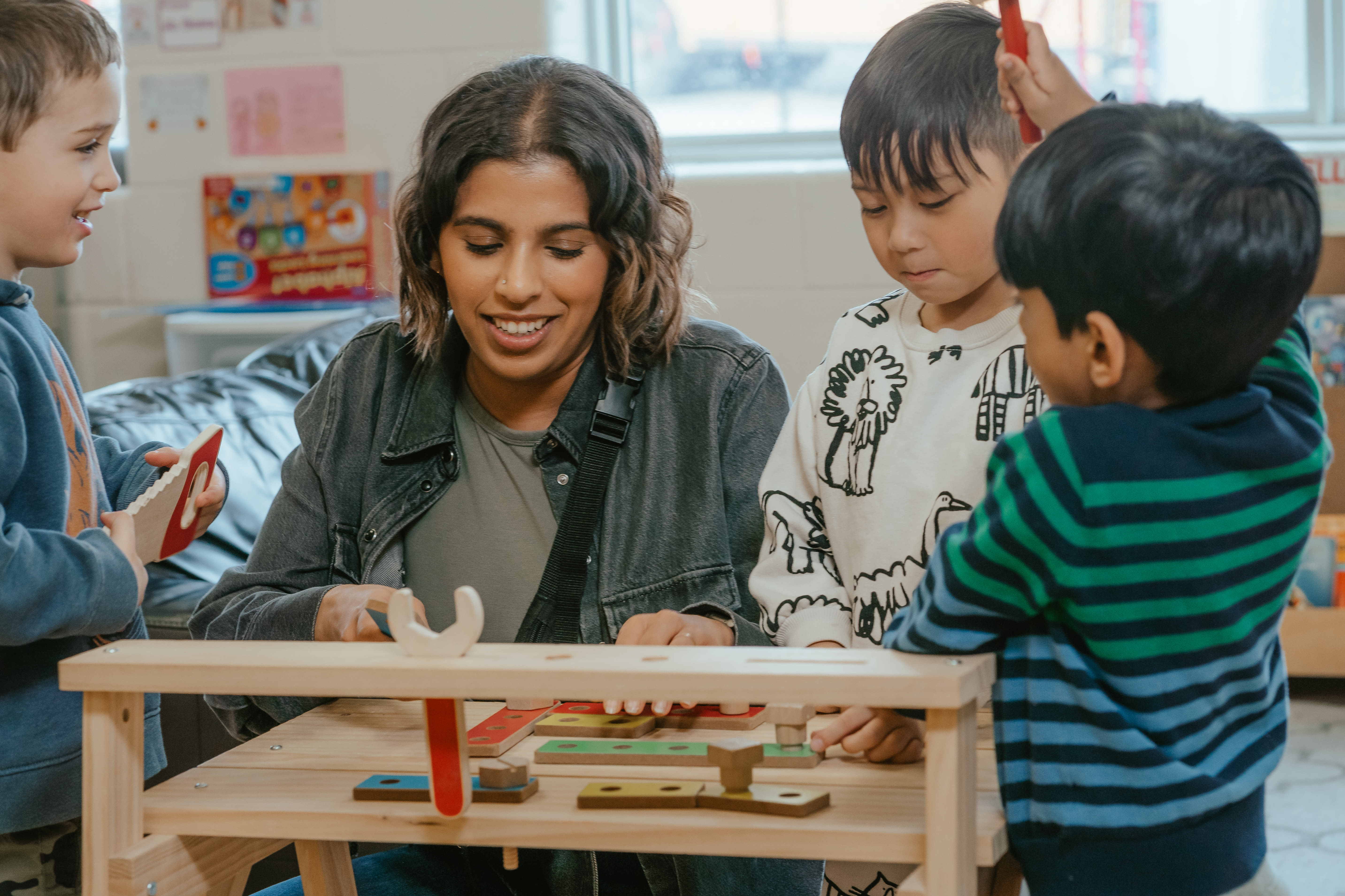 An educator plays with three children using a woodworking simulation setup in a YMCA child care classroom.