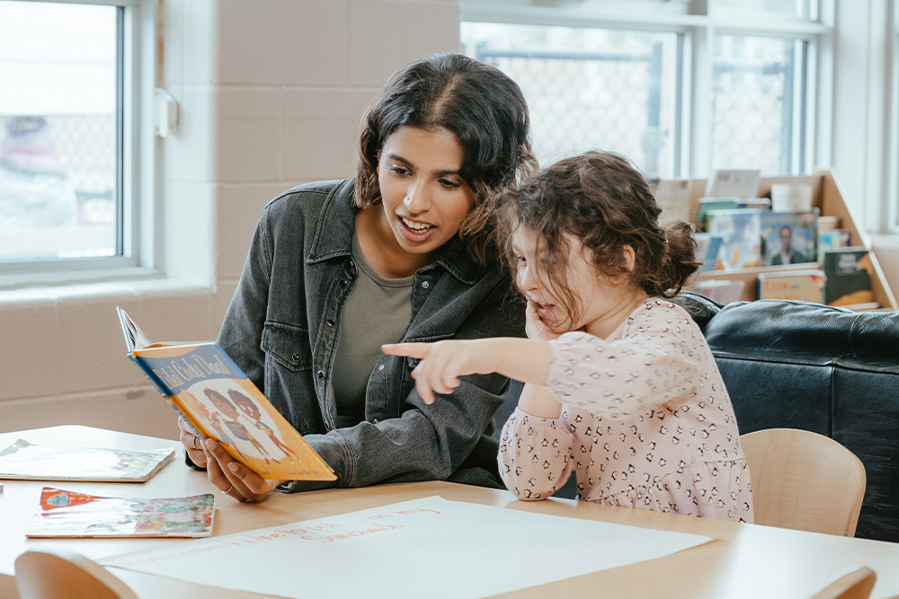 An educator in a grey jean jacket with short ombre hair reads a picture book titled "Bilaal Cooks Daal" with a child wearing a pink long-sleeve shirt who points to a photo in the book.