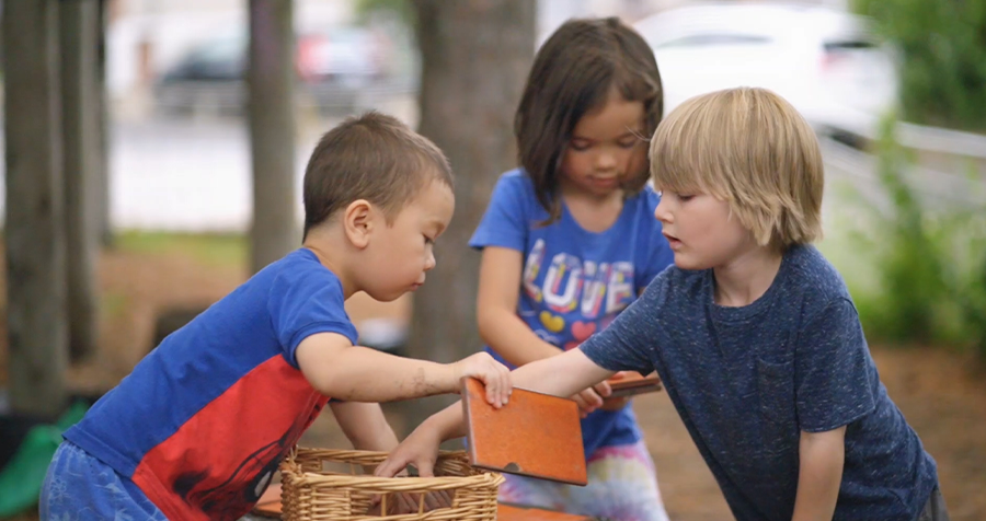  Three children, dressed in blue shirts, diligently lay tiles outdoors on the playground of a Licensed Summer School Break Child Care YMCA program. They carefully select the tiles from a rattan basket, engaged in the hands-on activity.