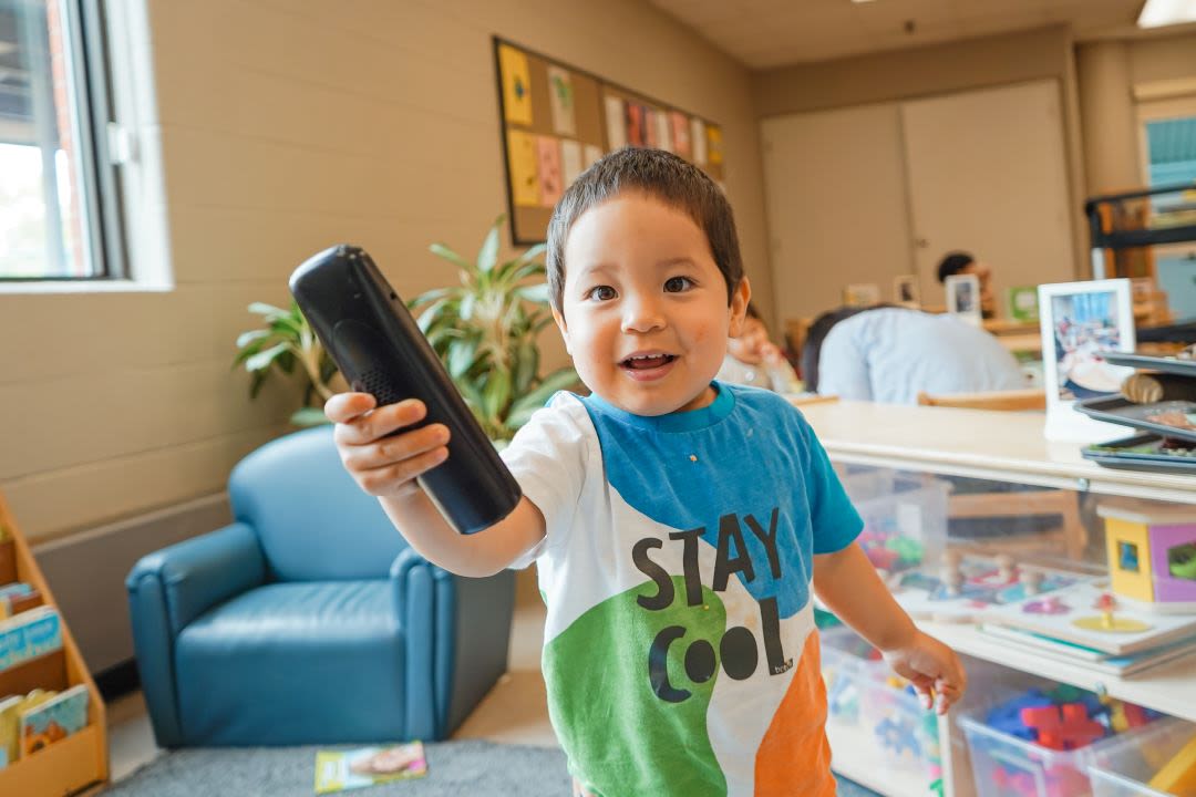 A child with brown hair holds a toy telephone in the play and reading area of an infant classroom, wearing a blue, white, and orange top that says "Stay Cool."