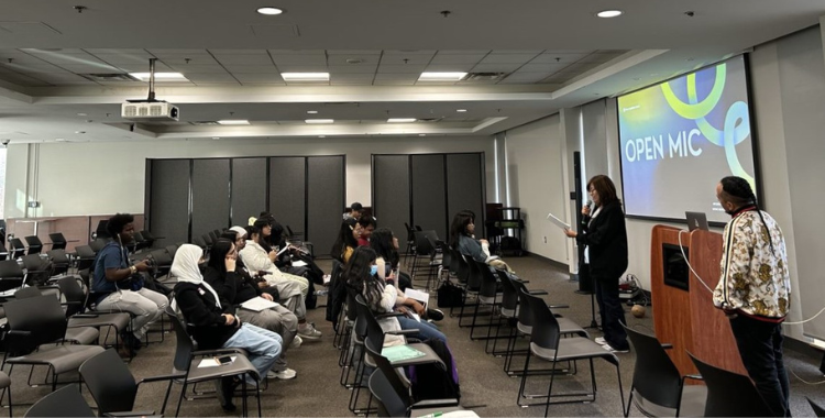 NYLD youth are sitting in chairs, facing a screen and podium, listening to a presenter and Beny Esguerra announce an open mic