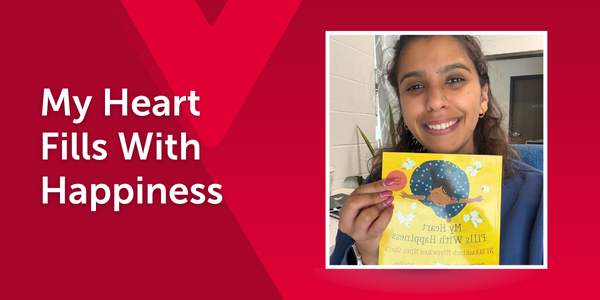 An educator smiles while holding My Heart Fills With Happiness by Monique Gray Smith. The cover features a joyful child. The image is overlaid on a red background with a faint Y M C A logo and the book title in text.