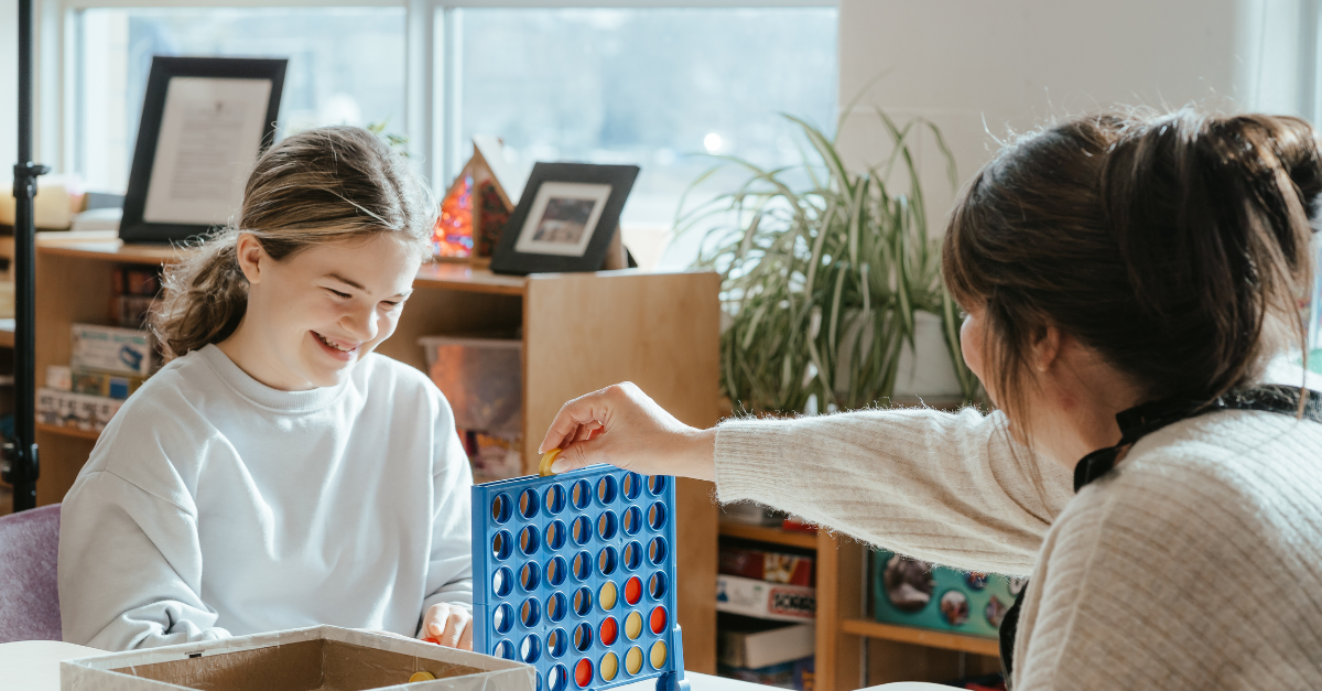 Child wearing a white sweater plays Connect Four with a YMCA educator in a before- and after-school classroom.