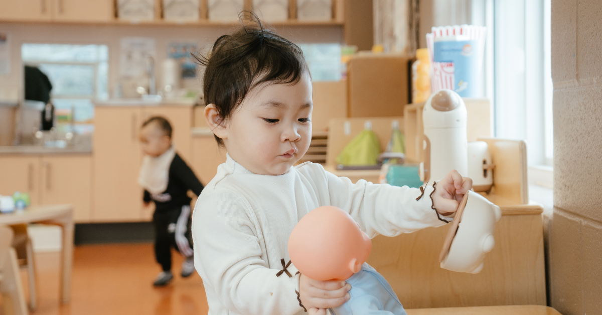 Child wearing a white long-sleeve shirt holds a toy baby in a blue outfit at a YMCA child care play area.