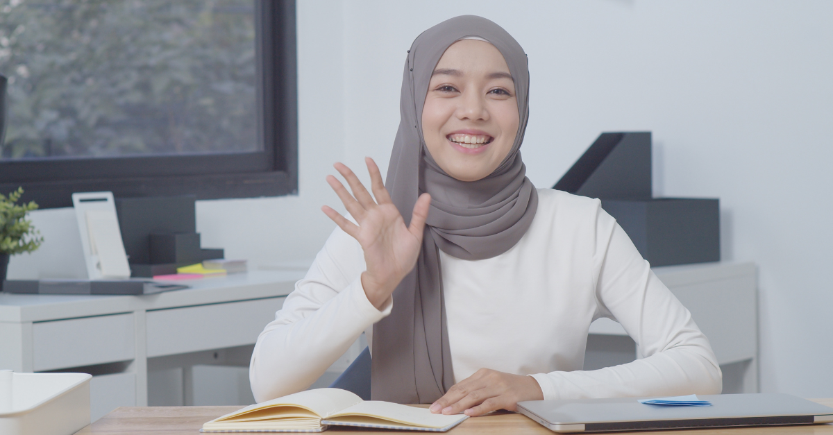 A person wearing a grey hijab and white long-sleeve shirt smiles and waves in a neutral office setting.
