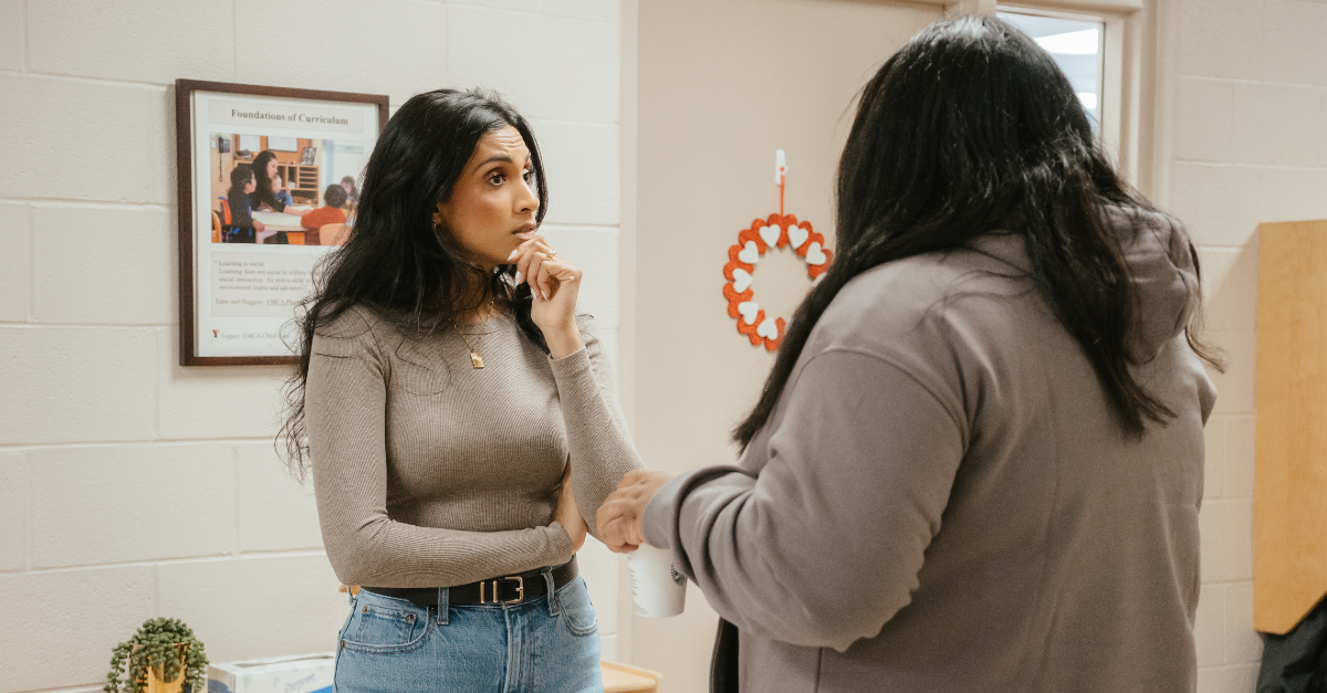 An educator in a beige long-sleeve shirt and blue jeans listens attentively to another educator in a grey hoodie.