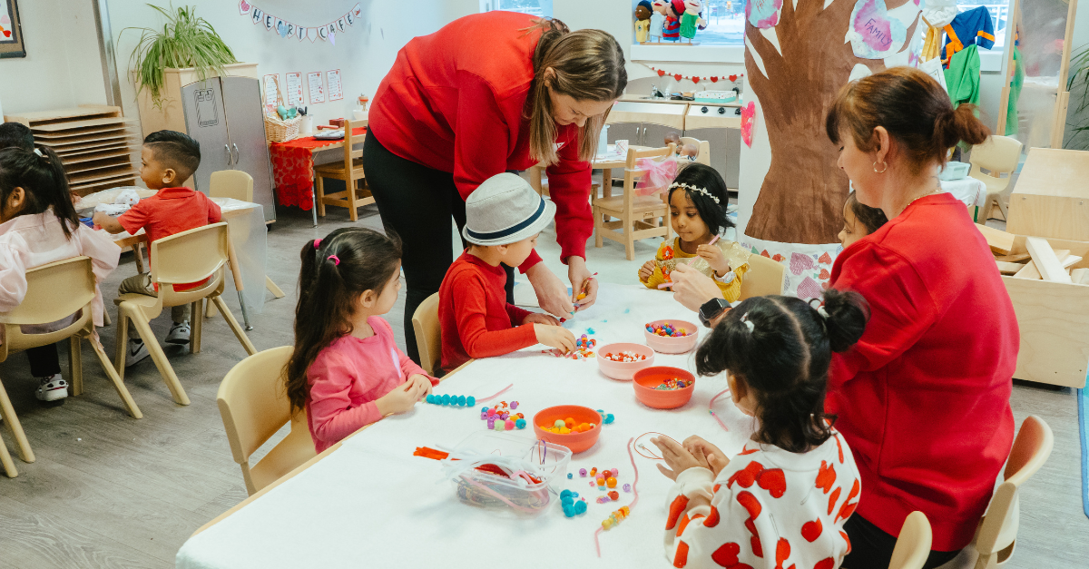 Two educators in red Y M C A Shine On crewnecks guide four children in a jewellery-making activity for Valentine's Day in a colourful Y M C A child care classroom.