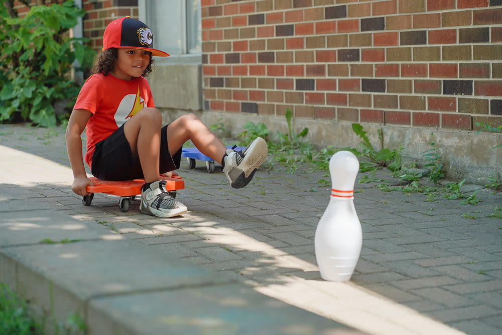 A child in a snapback hat and bright red top sits on a scooter board playing on an outdoor obstacle course with bowling pins at a YMCA child care playground.