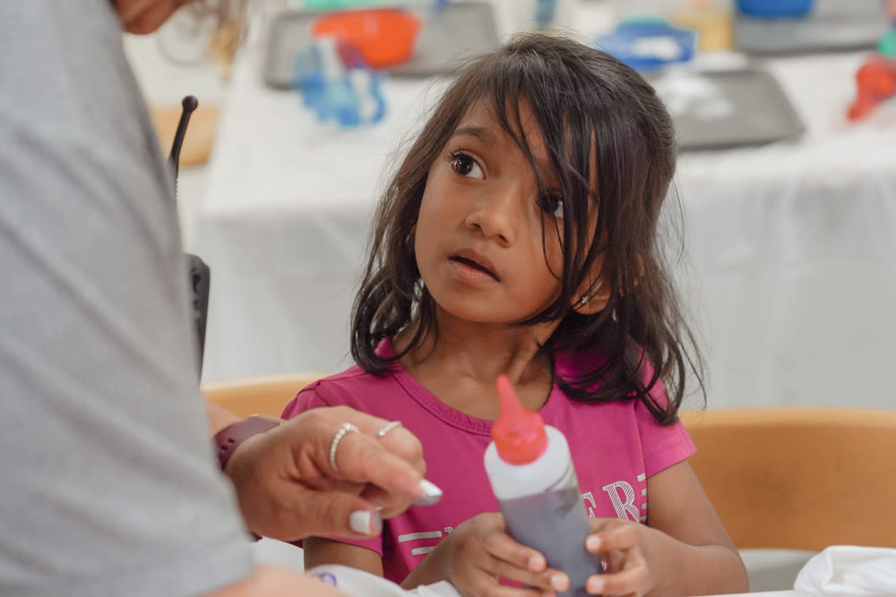 A curious child in a pink top looks up at an educator holding a tie-dye colour bottle in a YMCA child care classroom.