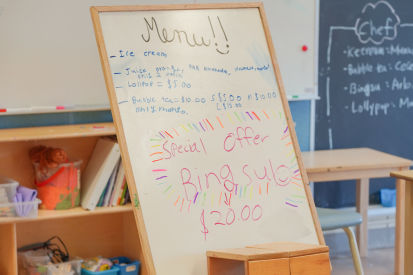 A whiteboard menu for the cafe reads: Ice cream, Juice: orange, pink lemonade, lemonade, mango, only 2 refills. Lollipop is $5. Bubble tea is $10 (only mango). Small is $5, Medium is $10, Large is $15. Special offer bingsu is $20 in a bigger red font.