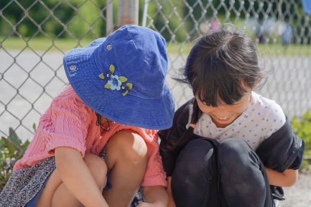 A child in a dark blue hat, pink top, and skirt plays next to a smiling child as they observe nature.