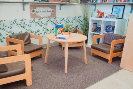 The reading corner of the classroom features three wooden chairs, a wooden table with two Harry Potter books, and a plant, along with a large collection of books displayed on multiple floating shelves.