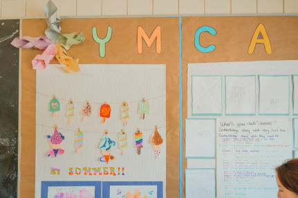 The wall in Jenny's classroom displays "Y.M.C.A" in colourful letters, surrounded by popsicle paper crafts created by the children to celebrate summer.