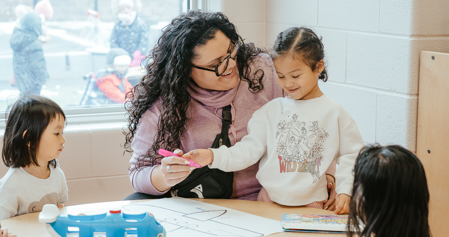 An educator in a pink hoodie with curly hair helps children colour an airplane drawing on a wooden desk in a child care classroom.