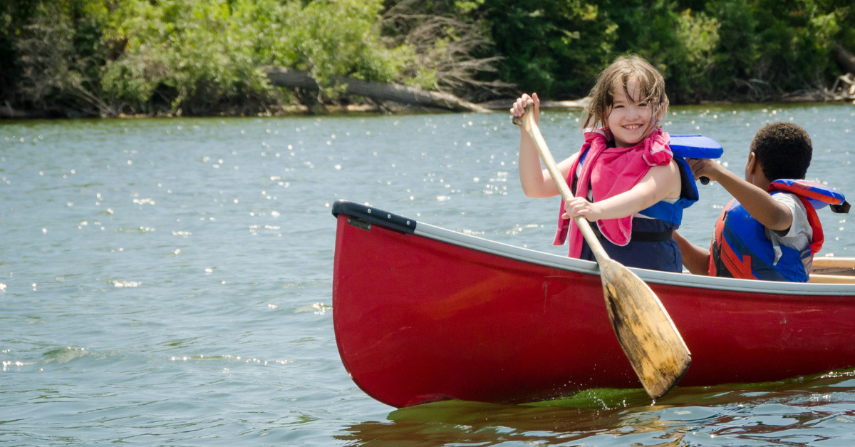 A child in a blue life vest and pink towel smiles while paddling a red canoe on a lake during a Y M C A camp program. Another child paddles in a separate canoe nearby.