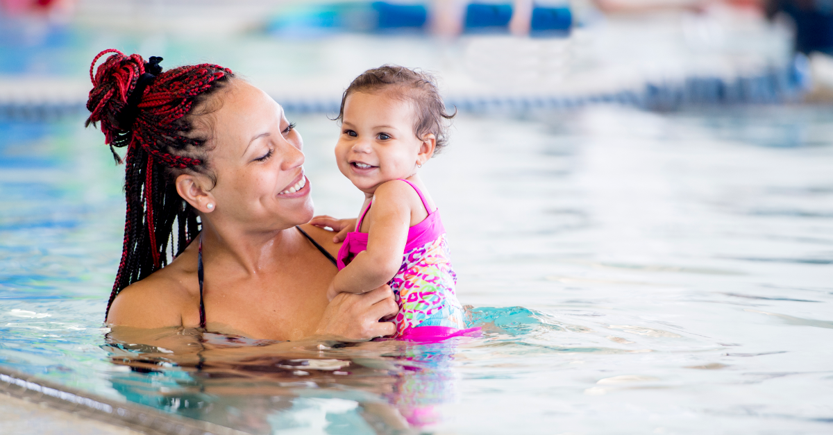 A parent with ombre braids, pinned up, holds their smiling toddler in a pink swimsuit at a Y M C A swimming pool. Both are smiling and enjoying the water together.