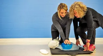 A person performs chest compressions on a dummy while a YMCA instructor supervises