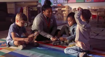 A child care educator sits on a carpet and plays with 3 children.