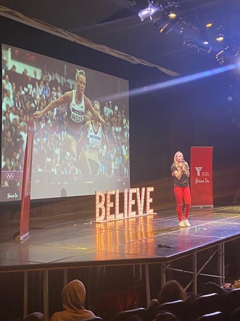 Sarah Wells presents on stage in front of a photo of her jumping a hurdle and next to the word "Believe" in lights.